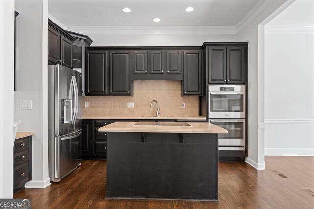 kitchen featuring an island with sink, light countertops, appliances with stainless steel finishes, ornamental molding, and dark wood-style flooring