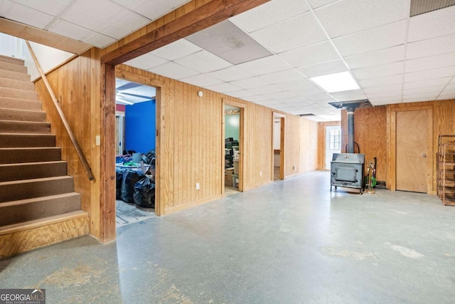 basement with a paneled ceiling, wooden walls, stairs, and a wood stove