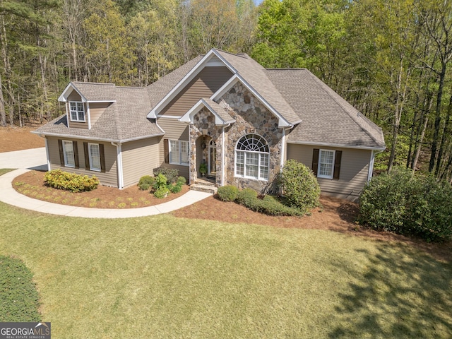 view of front of property featuring a front lawn, roof with shingles, a view of trees, and stone siding