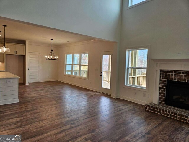 unfurnished living room featuring dark wood-style flooring, crown molding, a brick fireplace, baseboards, and a notable chandelier