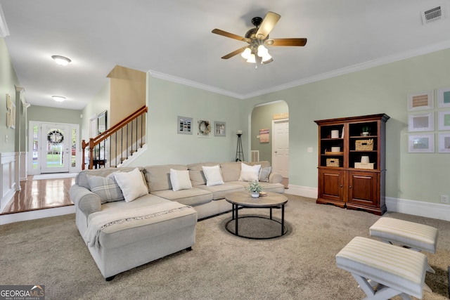carpeted living area with visible vents, arched walkways, ornamental molding, ceiling fan, and stairway