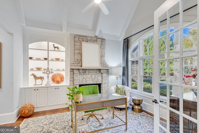 home office featuring baseboards, ceiling fan, a stone fireplace, vaulted ceiling with beams, and wood finished floors
