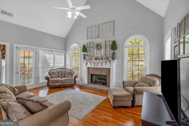 living room with high vaulted ceiling, visible vents, a brick fireplace, wood finished floors, and a ceiling fan