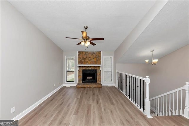 unfurnished living room featuring wood finished floors, a textured ceiling, a stone fireplace, baseboards, and ceiling fan with notable chandelier