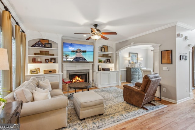 living area with a high end fireplace, crown molding, a ceiling fan, arched walkways, and light wood-style floors
