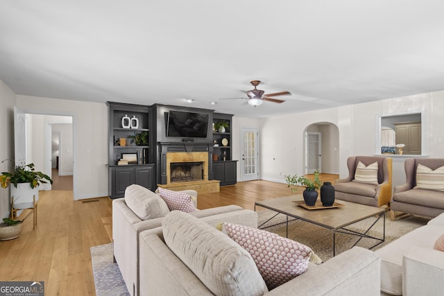 living area featuring light wood-style floors, arched walkways, a ceiling fan, a tile fireplace, and baseboards