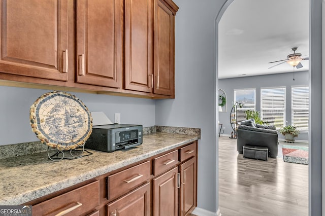 kitchen featuring wood finished floors, arched walkways, brown cabinets, ceiling fan, and baseboards