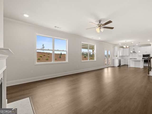 unfurnished living room with ceiling fan with notable chandelier, visible vents, crown molding, and dark wood-style floors
