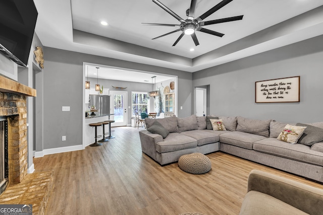 living room featuring a tray ceiling, wood finished floors, a fireplace with raised hearth, baseboards, and ceiling fan with notable chandelier