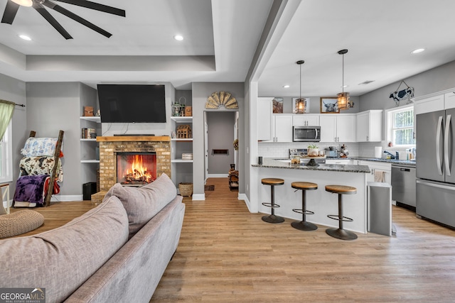 living room featuring baseboards, light wood-style floors, a ceiling fan, and a brick fireplace