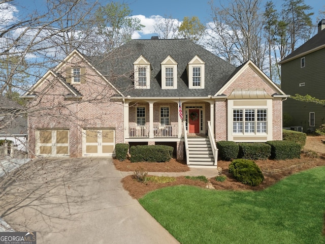 new england style home with a garage, driveway, brick siding, a porch, and a shingled roof