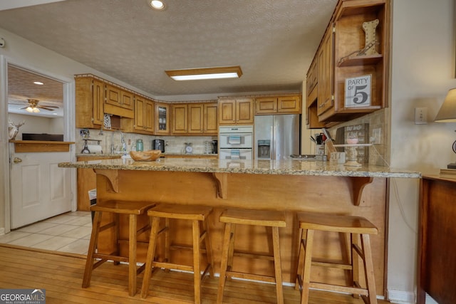 kitchen featuring light stone counters, backsplash, stainless steel fridge, a peninsula, and brown cabinetry