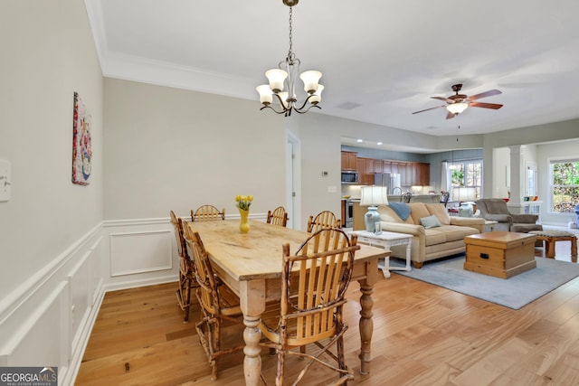 dining space with crown molding, light wood-type flooring, a wainscoted wall, ceiling fan with notable chandelier, and a decorative wall