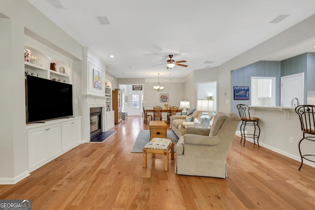 living room with light wood finished floors, a glass covered fireplace, built in features, ceiling fan with notable chandelier, and baseboards