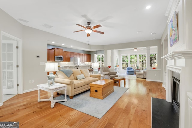 living area with visible vents, a fireplace with flush hearth, ornate columns, light wood-type flooring, and a ceiling fan
