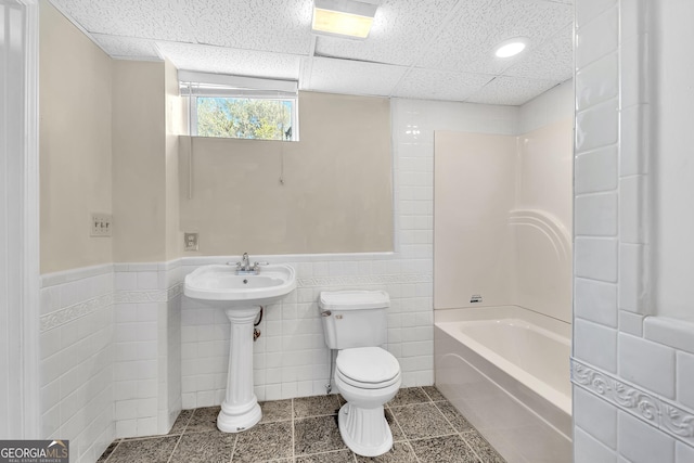bathroom with toilet, tile walls, a paneled ceiling, tub / shower combination, and wainscoting