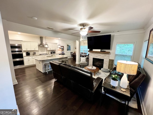living area featuring a glass covered fireplace, ornamental molding, a ceiling fan, dark wood-type flooring, and visible vents