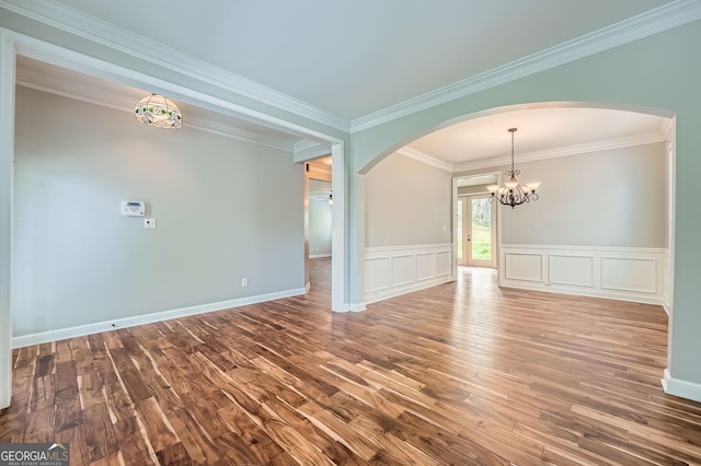 empty room with wood finished floors, a chandelier, arched walkways, a wainscoted wall, and ornamental molding