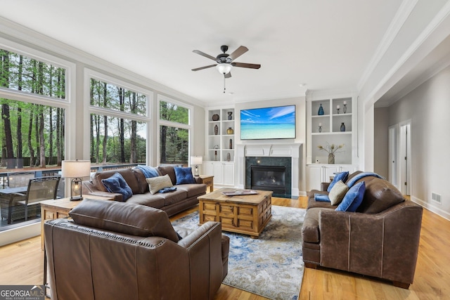 living room featuring crown molding, ceiling fan, a fireplace, light wood-style flooring, and built in shelves