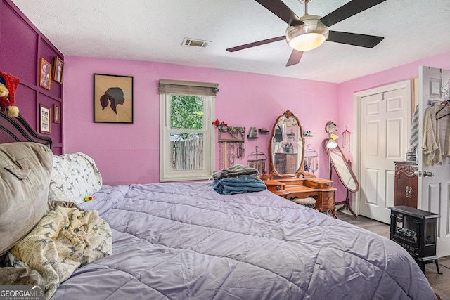 bedroom with wood finished floors, a textured ceiling, a wood stove, and ceiling fan