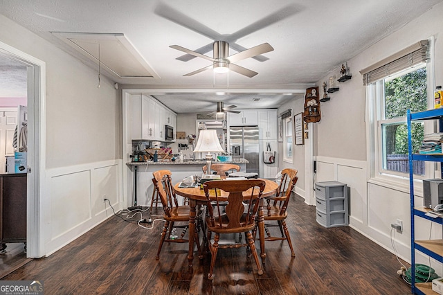 dining room featuring attic access, a decorative wall, dark wood-style flooring, a ceiling fan, and wainscoting