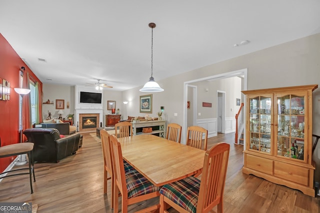dining space featuring a glass covered fireplace, light wood-style flooring, a wainscoted wall, and ceiling fan