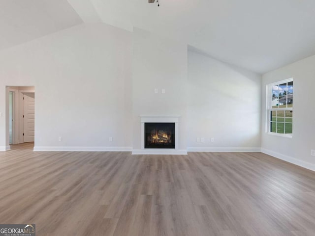 unfurnished living room featuring baseboards, high vaulted ceiling, light wood-style floors, and a lit fireplace