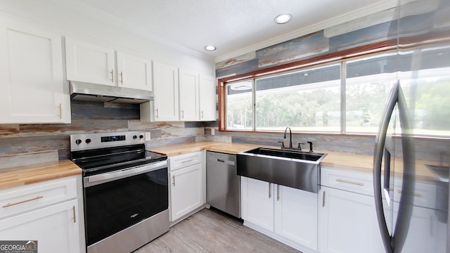 kitchen with appliances with stainless steel finishes, butcher block countertops, under cabinet range hood, ornamental molding, and a sink