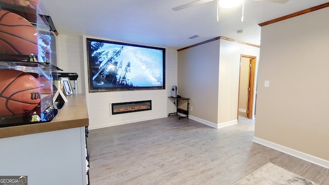 home theater with light wood finished floors, a ceiling fan, a glass covered fireplace, baseboards, and ornamental molding