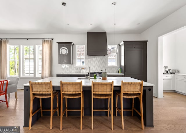 kitchen featuring custom range hood, light stone countertops, a kitchen island with sink, decorative backsplash, and a sink