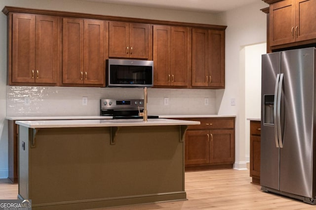 kitchen with appliances with stainless steel finishes, a kitchen island with sink, light countertops, light wood finished floors, and backsplash