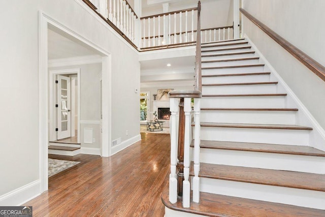 stairs with crown molding, wood finished floors, a warm lit fireplace, a towering ceiling, and baseboards