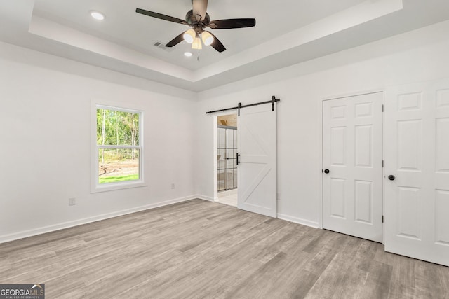 unfurnished bedroom with baseboards, a barn door, light wood-style floors, a tray ceiling, and recessed lighting
