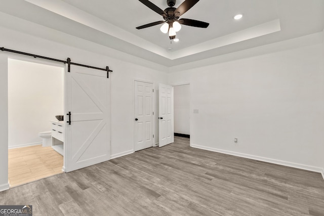 unfurnished bedroom featuring baseboards, a barn door, a tray ceiling, recessed lighting, and wood finished floors