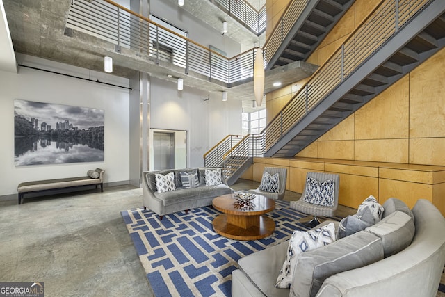 living room featuring concrete floors, baseboards, wooden walls, a high ceiling, and stairs