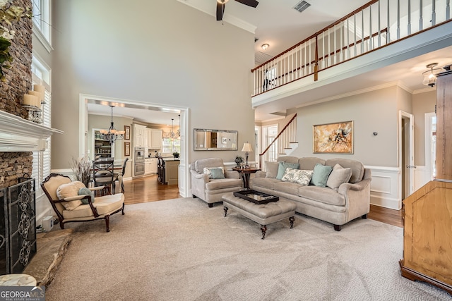 living area featuring ornamental molding, a stone fireplace, a healthy amount of sunlight, ceiling fan with notable chandelier, and a wainscoted wall