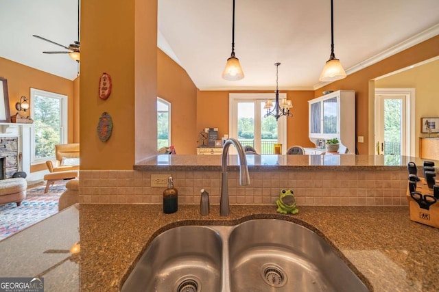 kitchen with a sink, ornamental molding, dark stone counters, ceiling fan with notable chandelier, and pendant lighting
