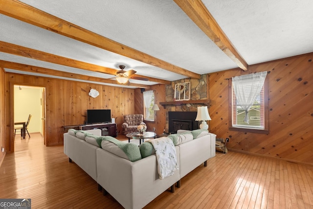 living area featuring a stone fireplace, ceiling fan, a textured ceiling, beamed ceiling, and light wood finished floors