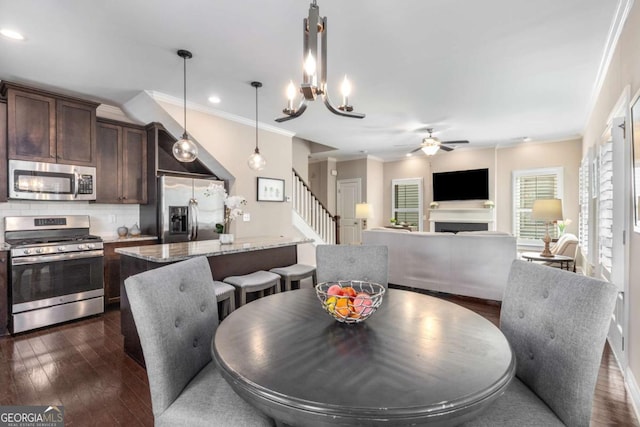 dining space featuring ornamental molding, a fireplace, stairway, dark wood-style floors, and ceiling fan with notable chandelier