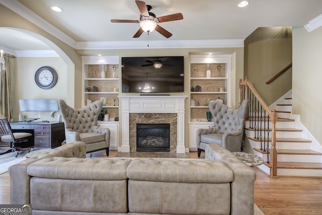 living area with built in shelves, a ceiling fan, crown molding, wood finished floors, and stairs