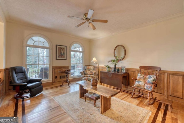 living area featuring a wainscoted wall, a ceiling fan, light wood-style flooring, ornamental molding, and a textured ceiling