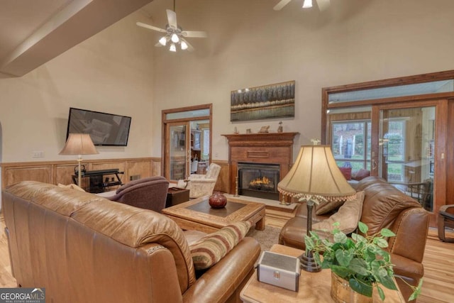 living area featuring a warm lit fireplace, ceiling fan, high vaulted ceiling, wood finished floors, and wainscoting