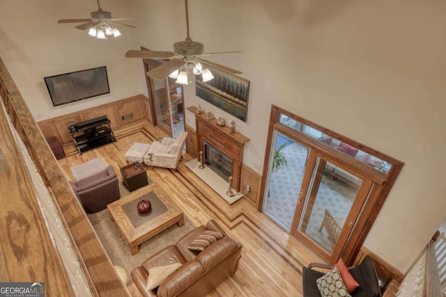 living room featuring a fireplace with raised hearth, a towering ceiling, a ceiling fan, light wood-style flooring, and a wainscoted wall