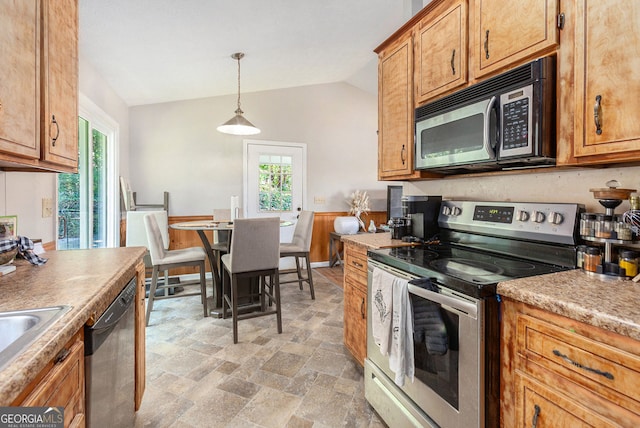 kitchen with stone finish floor, vaulted ceiling, stainless steel appliances, hanging light fixtures, and brown cabinets