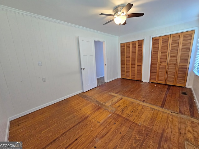 unfurnished bedroom featuring ornamental molding, a ceiling fan, hardwood / wood-style floors, multiple closets, and baseboards