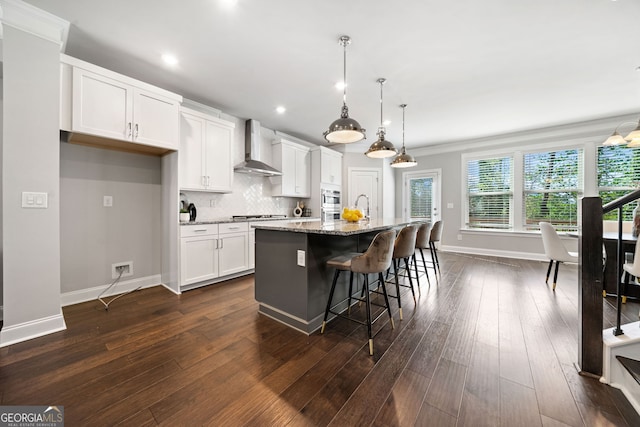 kitchen featuring wall chimney exhaust hood, tasteful backsplash, white cabinetry, dark wood-style flooring, and a center island with sink