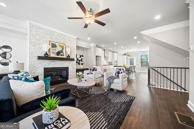 living area with a stone fireplace, ornamental molding, dark wood-type flooring, visible vents, and ceiling fan