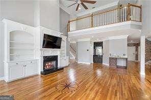 unfurnished living room featuring a ceiling fan, crown molding, wood finished floors, a towering ceiling, and a lit fireplace