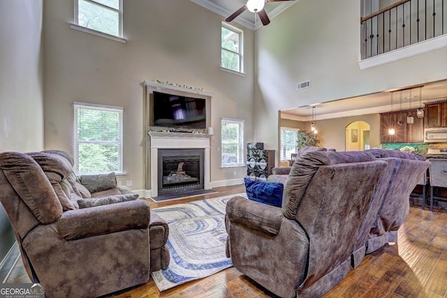 living area with visible vents, a ceiling fan, crown molding, a towering ceiling, and a glass covered fireplace