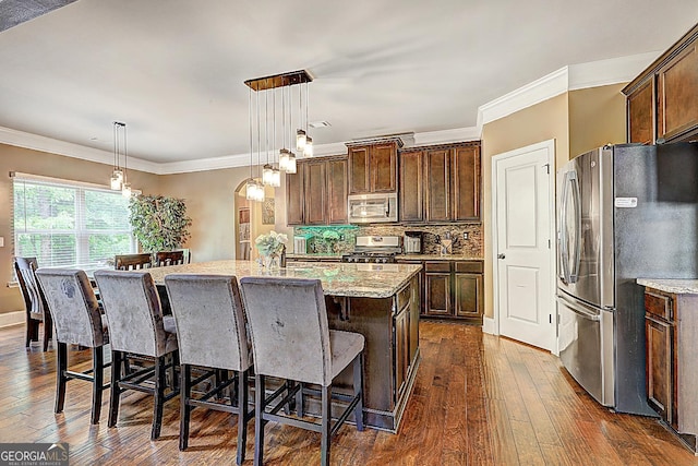 kitchen with appliances with stainless steel finishes, an island with sink, light stone countertops, and dark wood-style flooring